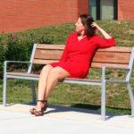 A woman in a red dress sits on a wooden bench outside near grass and flowers, looking to her left on a sunny day.