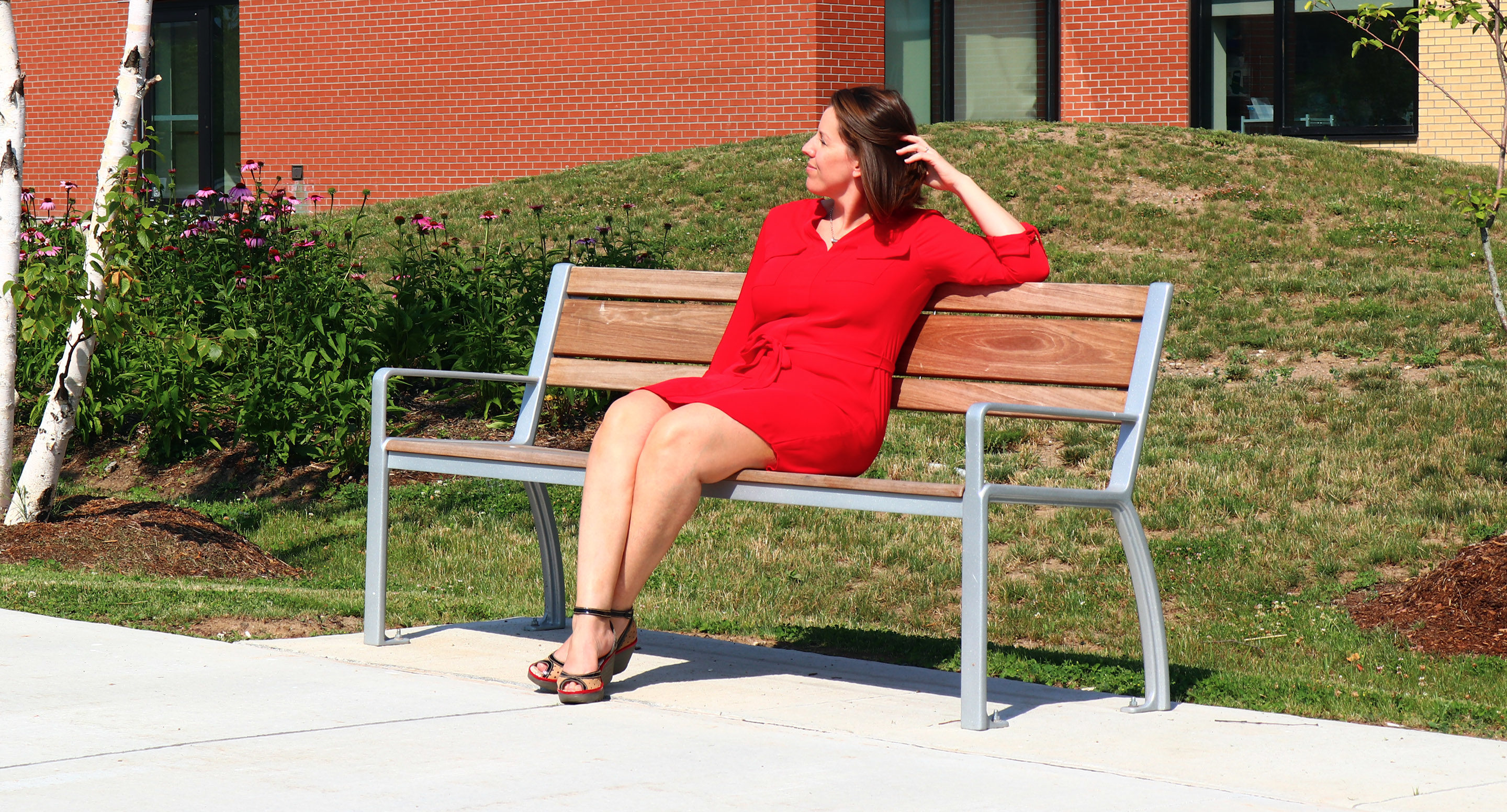 A woman in a red dress sits on a wooden bench outside near grass and flowers, looking to her left on a sunny day.