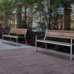 Three empty wooden benches with metal frames are placed along a paved walkway outside a building with large windows and greenery.