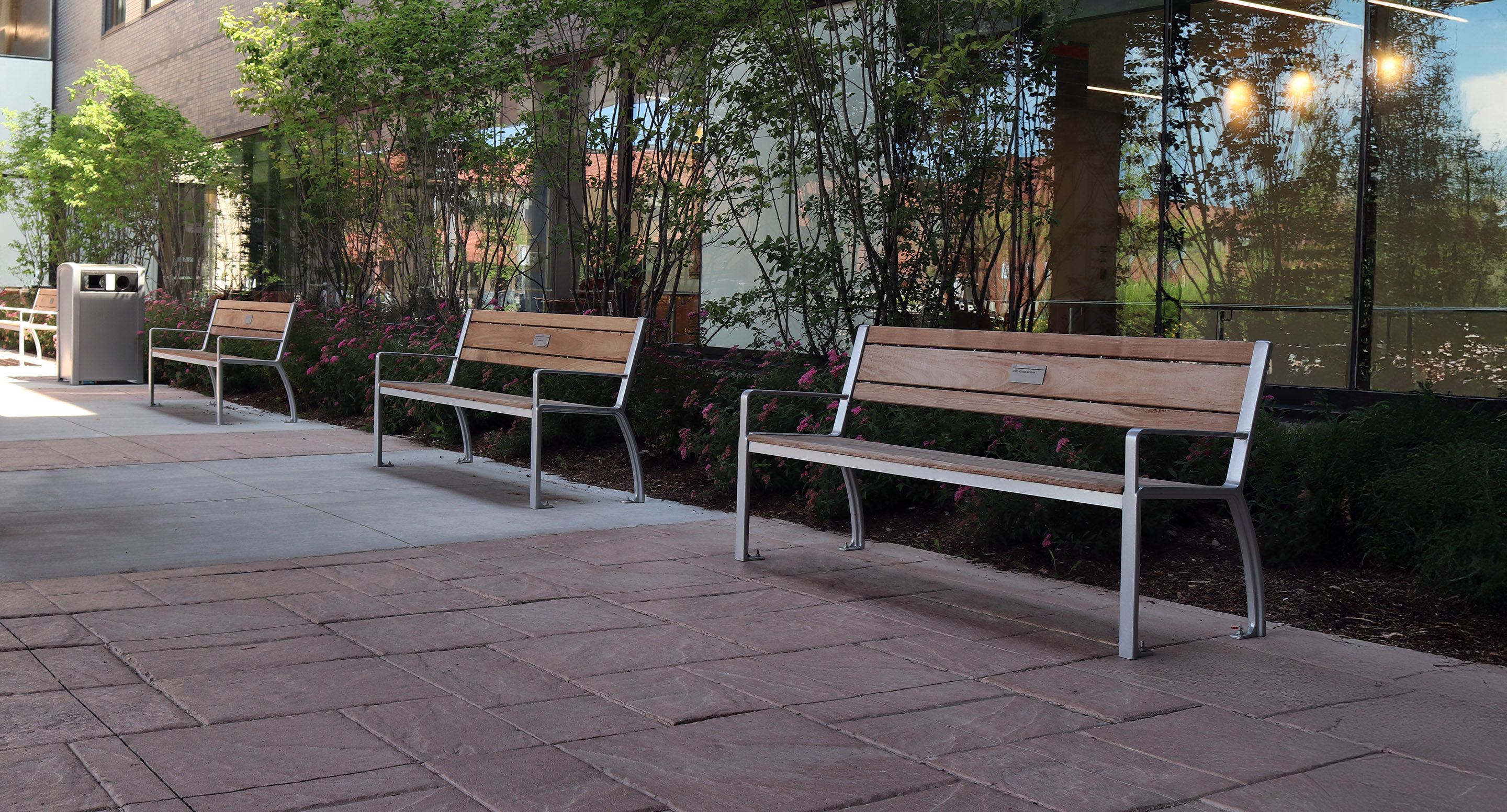 Three empty wooden benches with metal frames are placed along a paved walkway outside a building with large windows and greenery.