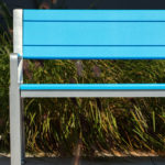 A bright blue metal bench with a silver frame is placed in front of tall ornamental grass in an outdoor setting.