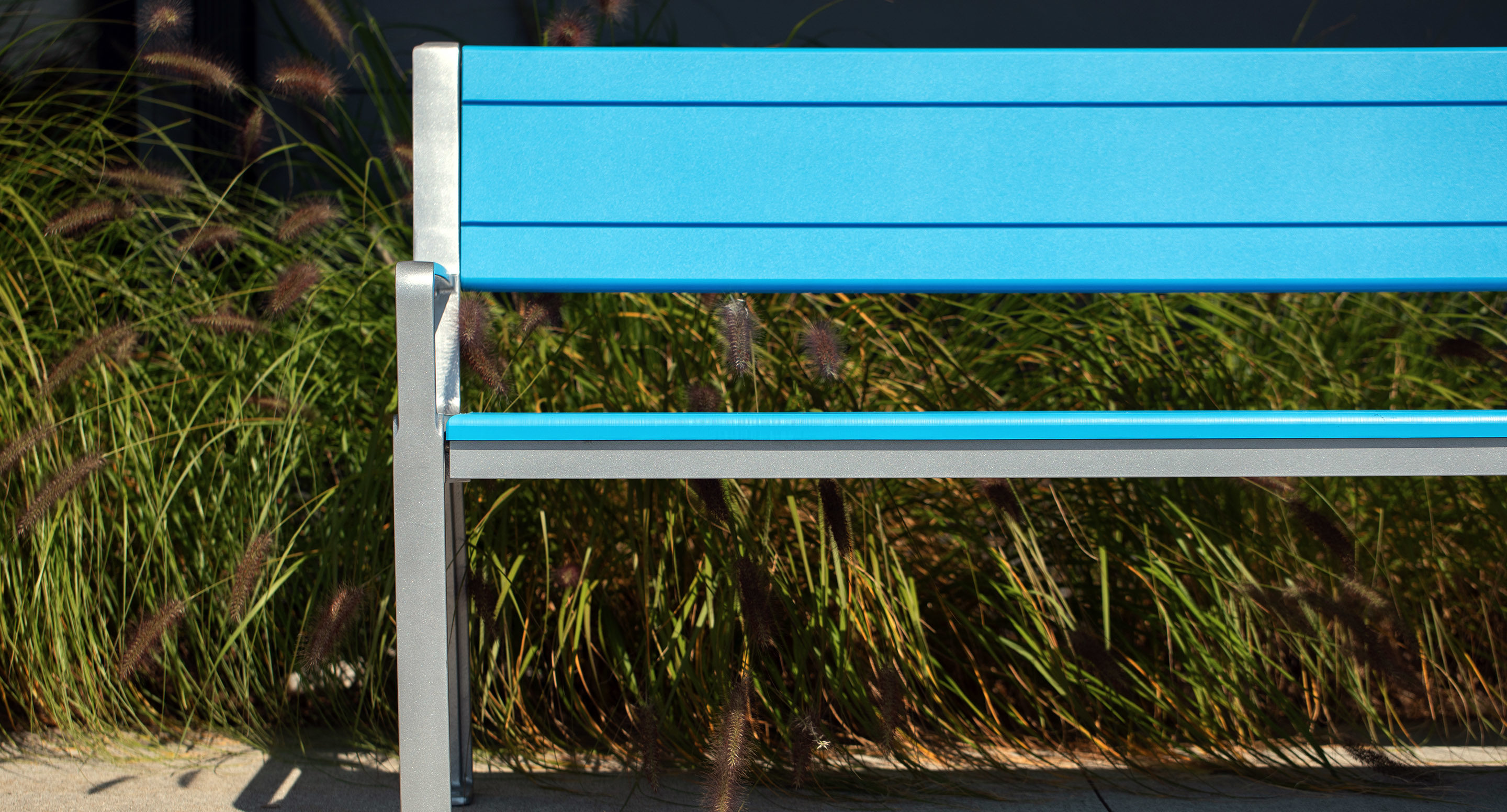 A bright blue metal bench with a silver frame is placed in front of tall ornamental grass in an outdoor setting.