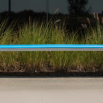 A modern metal bench with a blue seat is placed on a concrete surface in front of tall ornamental grasses.