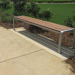 A modern wooden bench with metal legs is placed beside a concrete sidewalk, bordered by plants and mulch, overlooking a grassy area.