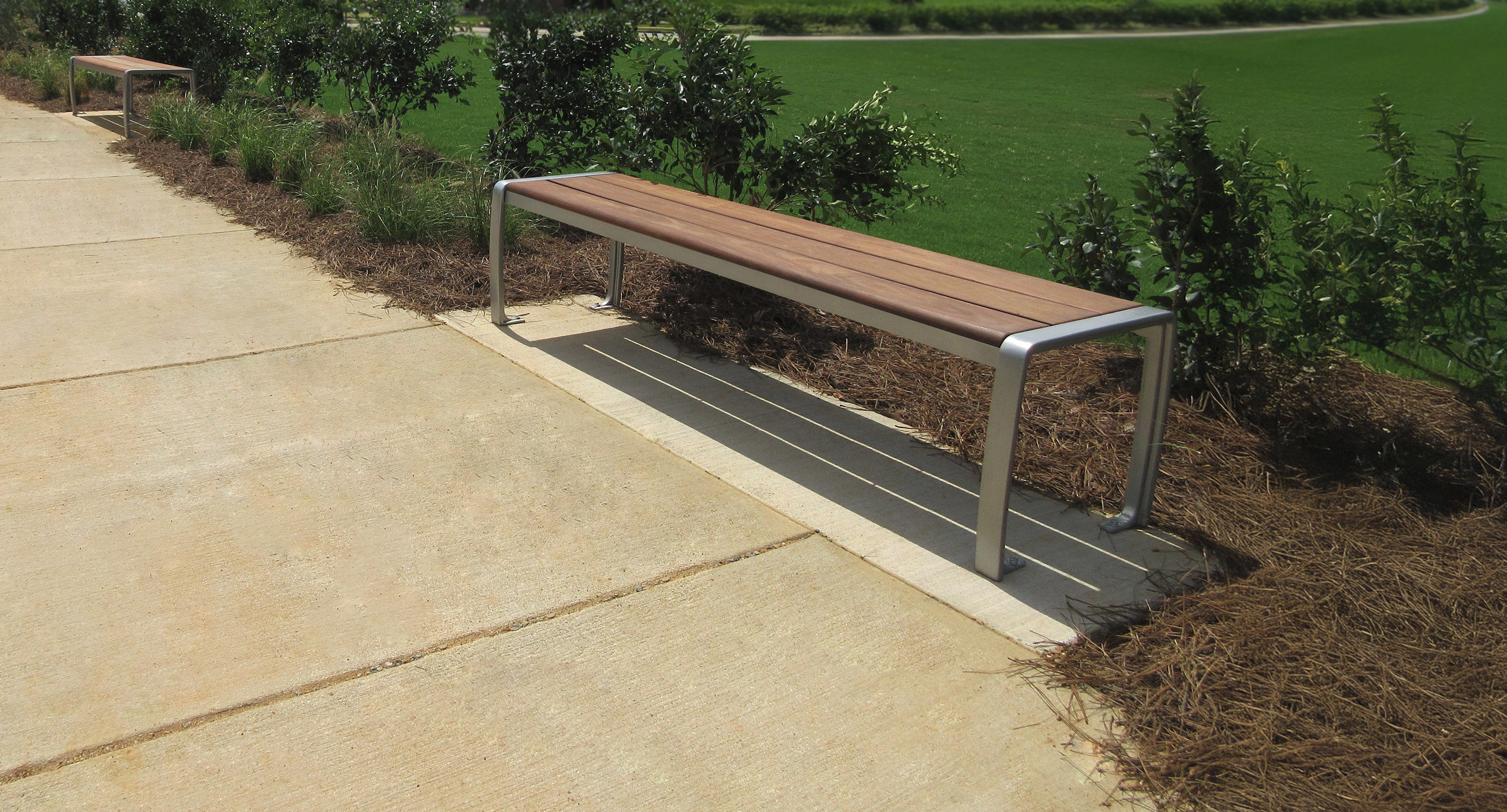 A modern wooden bench with metal legs is placed beside a concrete sidewalk, bordered by plants and mulch, overlooking a grassy area.