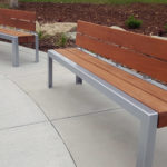 Two modern wooden benches with metal frames are placed on a curved concrete walkway in an outdoor landscaped area.