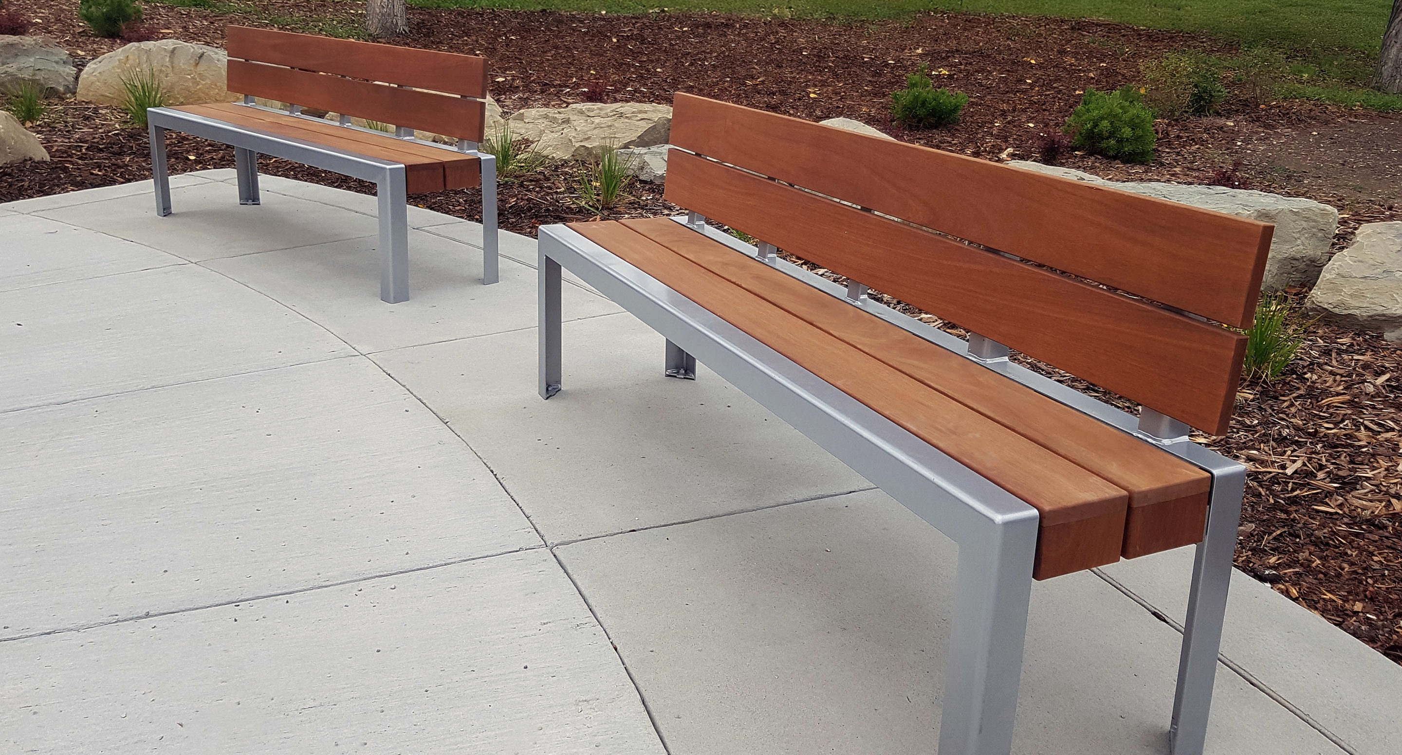 maglin_mbe-1050-backed-benches-1 Two modern wooden benches with metal frames are placed on a curved concrete walkway in an outdoor landscaped area.