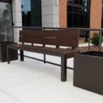 A brown metal bench with planters on both sides is placed on a sidewalk outside a modern building with glass doors and brick accents.