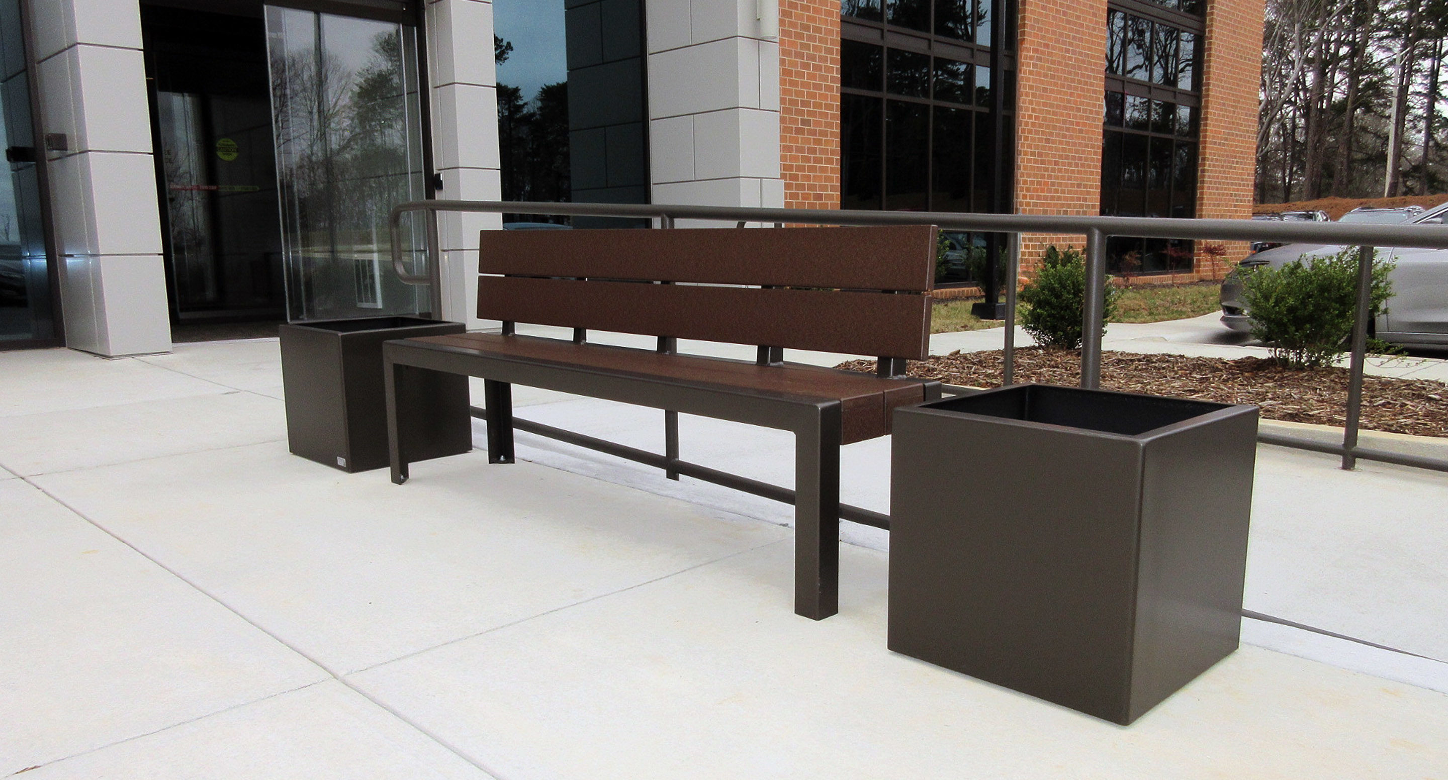 maglin_mbe-1050-backed-benches-2 A brown metal bench with planters on both sides is placed on a sidewalk outside a modern building with glass doors and brick accents.