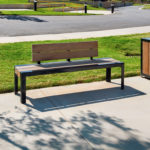 A modern bench with a wooden backrest and a matching trash bin are placed on a concrete sidewalk next to a grassy area in a suburban outdoor setting.