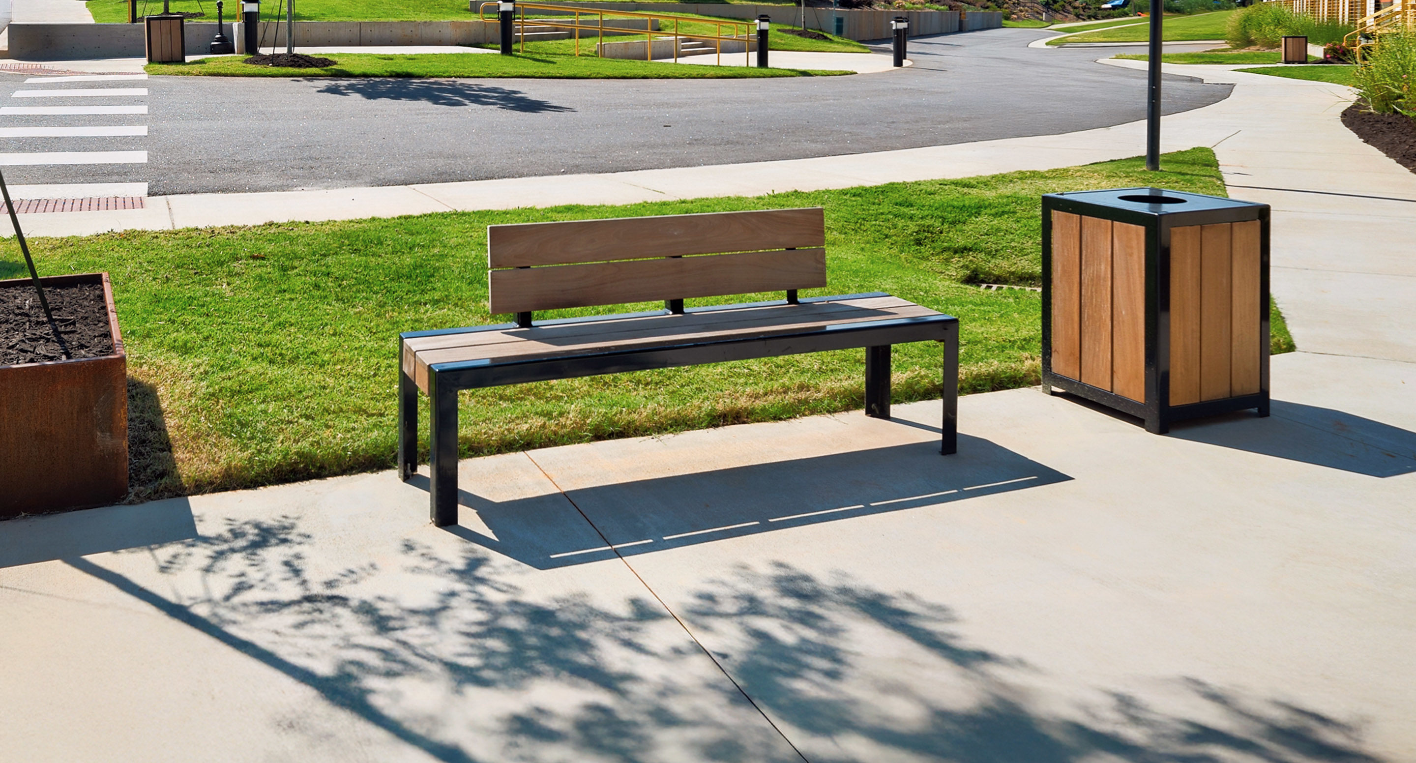 maglin_mbe-1050-backed-benches-3 A modern bench with a wooden backrest and a matching trash bin are placed on a concrete sidewalk next to a grassy area in a suburban outdoor setting.
