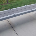 A modern gray metal bench with arm dividers is positioned on a concrete sidewalk next to a grassy area.
