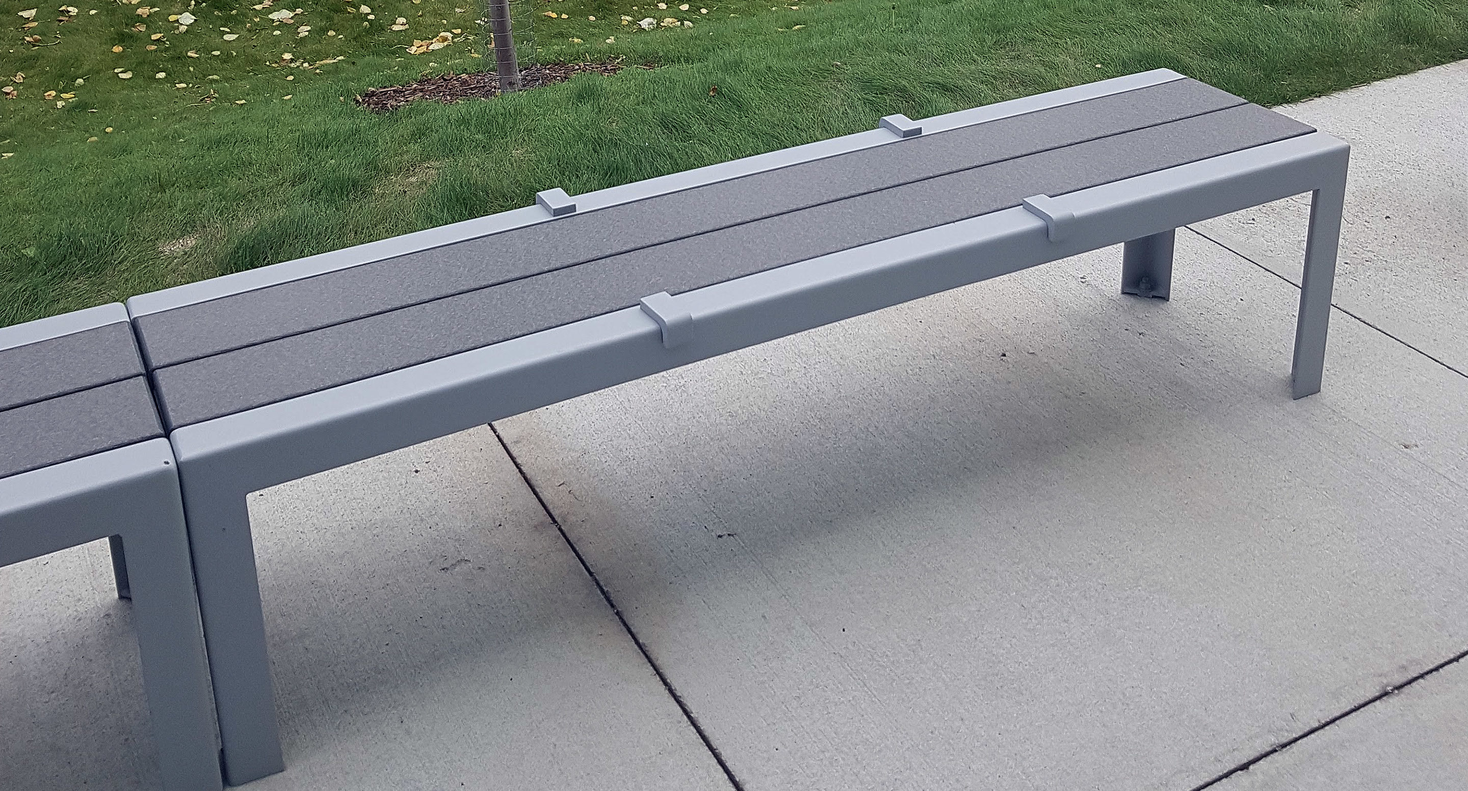 maglin_mbe-1050-backless-benches-1 A modern gray metal bench with arm dividers is positioned on a concrete sidewalk next to a grassy area.