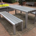 A modern outdoor seating area with a rectangular metal table and three matching benches on a brick patio, near a raised flower bed with yellow flowers.