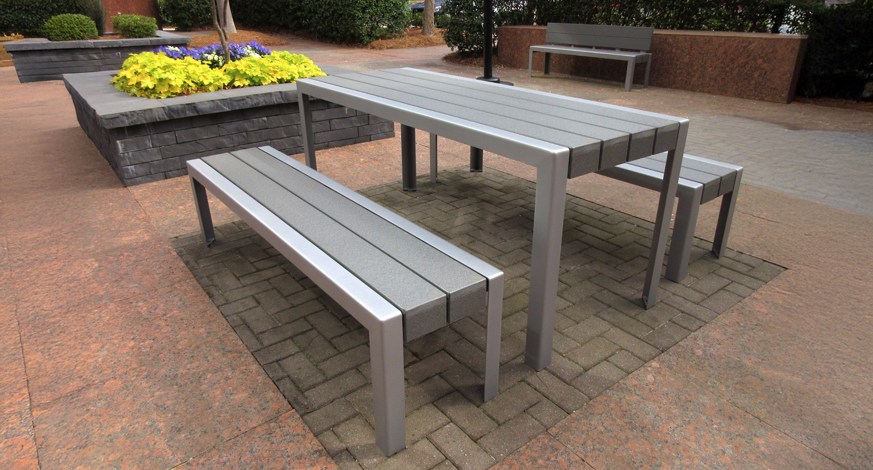 maglin_mbe-1050-backless-benches-2 A modern outdoor seating area with a rectangular metal table and three matching benches on a brick patio, near a raised flower bed with yellow flowers.