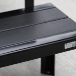 A close-up view of a black metal bench with a shiny finish, featuring a small manufacturer's label on one corner, placed on a light-colored surface.