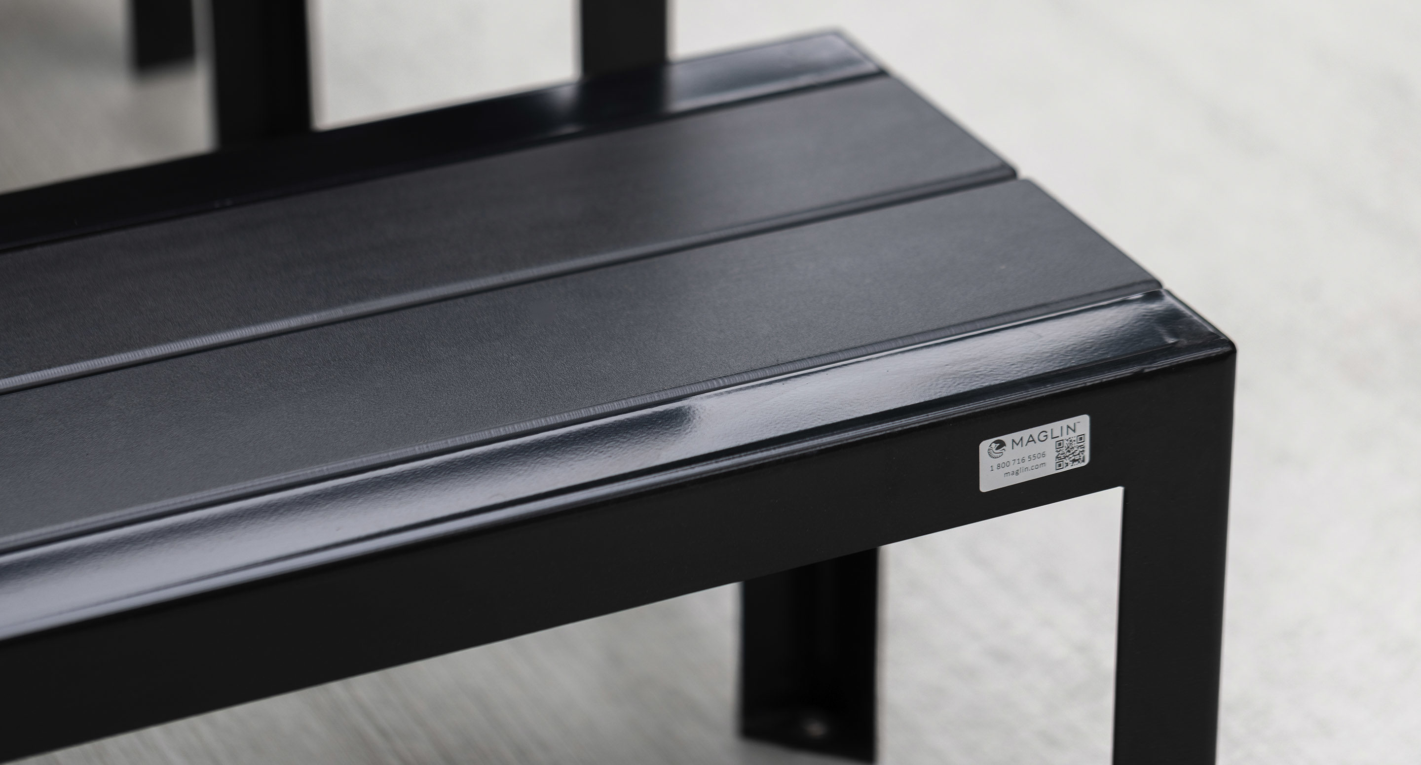 maglin_mbe-1050-backless-benches-3 A close-up view of a black metal bench with a shiny finish, featuring a small manufacturer's label on one corner, placed on a light-colored surface.