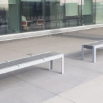 Deux bancs modernes en métal avec des barres de séparation sont placés sur un patio en béton à l'extérieur d'un bâtiment avec de grandes baies vitrées.