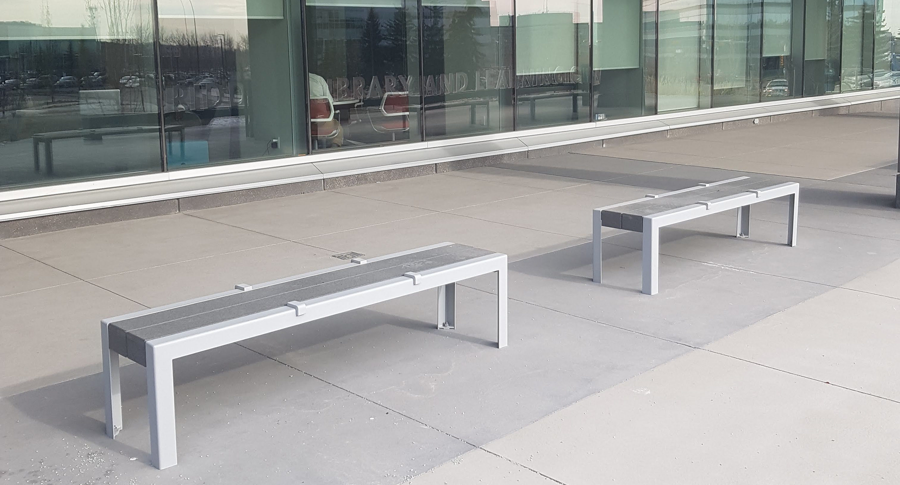 maglin_mbe-1050-bench-skate-deterrents-1 Deux bancs modernes en métal avec des barres de séparation sont placés sur un patio en béton à l'extérieur d'un bâtiment avec de grandes baies vitrées.