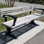 Un banc public en bois avec des accoudoirs et des pieds en métal noir est placé sur une dalle de béton à côté d'un chemin pavé dans un espace extérieur herbeux.