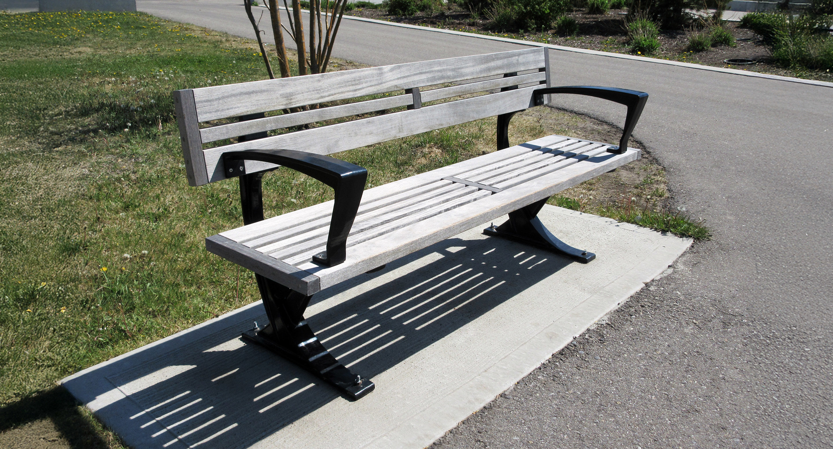 Un banc public en bois avec des accoudoirs et des pieds en métal noir est placé sur une dalle de béton à côté d'un chemin pavé dans un espace extérieur herbeux.
