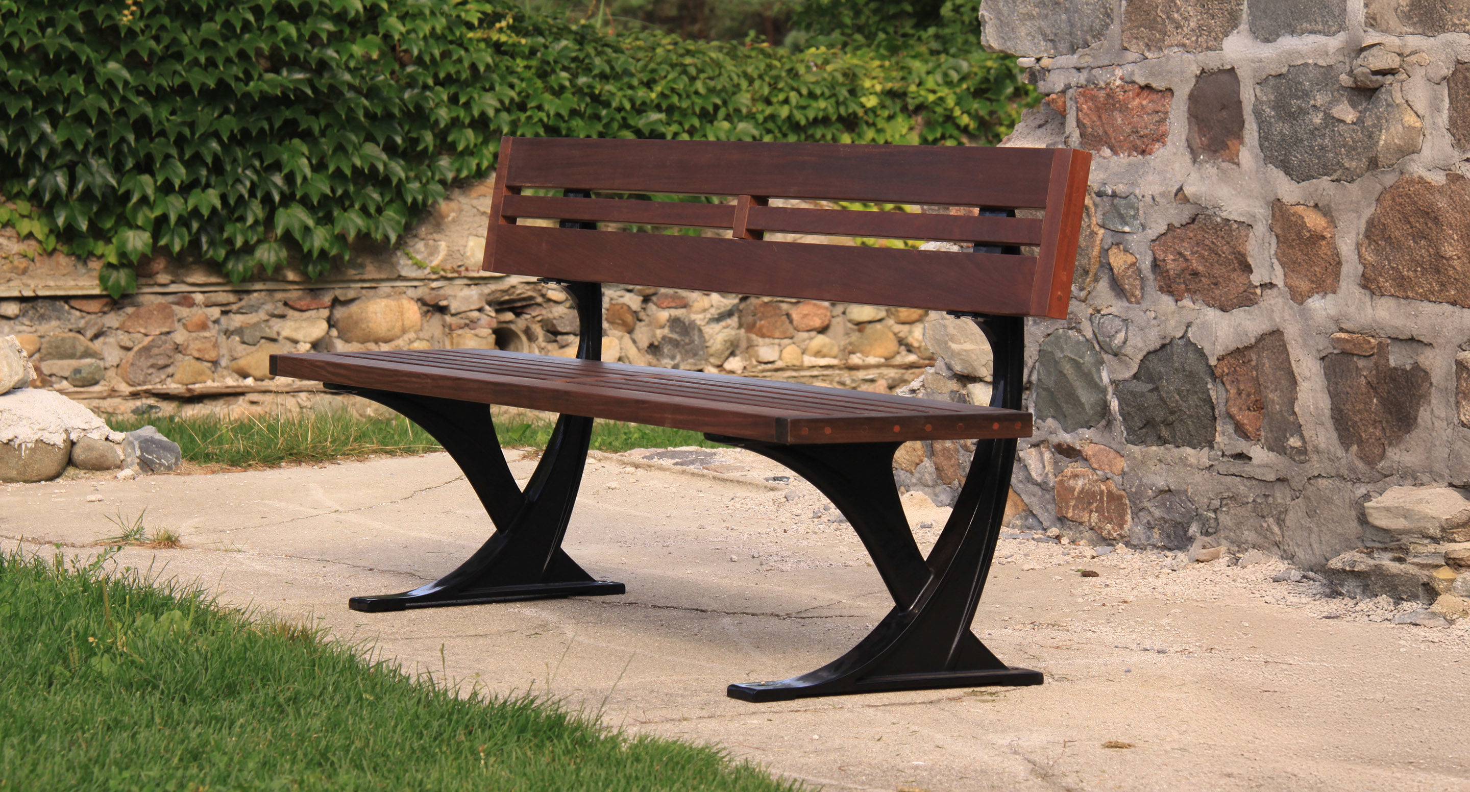 maglin_mbe-1200-backed-benches-2 Un banc en bois avec une armature métallique est placé sur un chemin en béton à côté d'un mur en pierre et d'un carré d'herbe.
