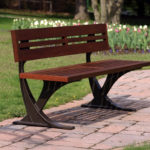 Un banc public en bois avec un cadre en métal est placé sur un sentier en pierre dans un parc herbeux avec des arbres et des fleurs en arrière-plan.