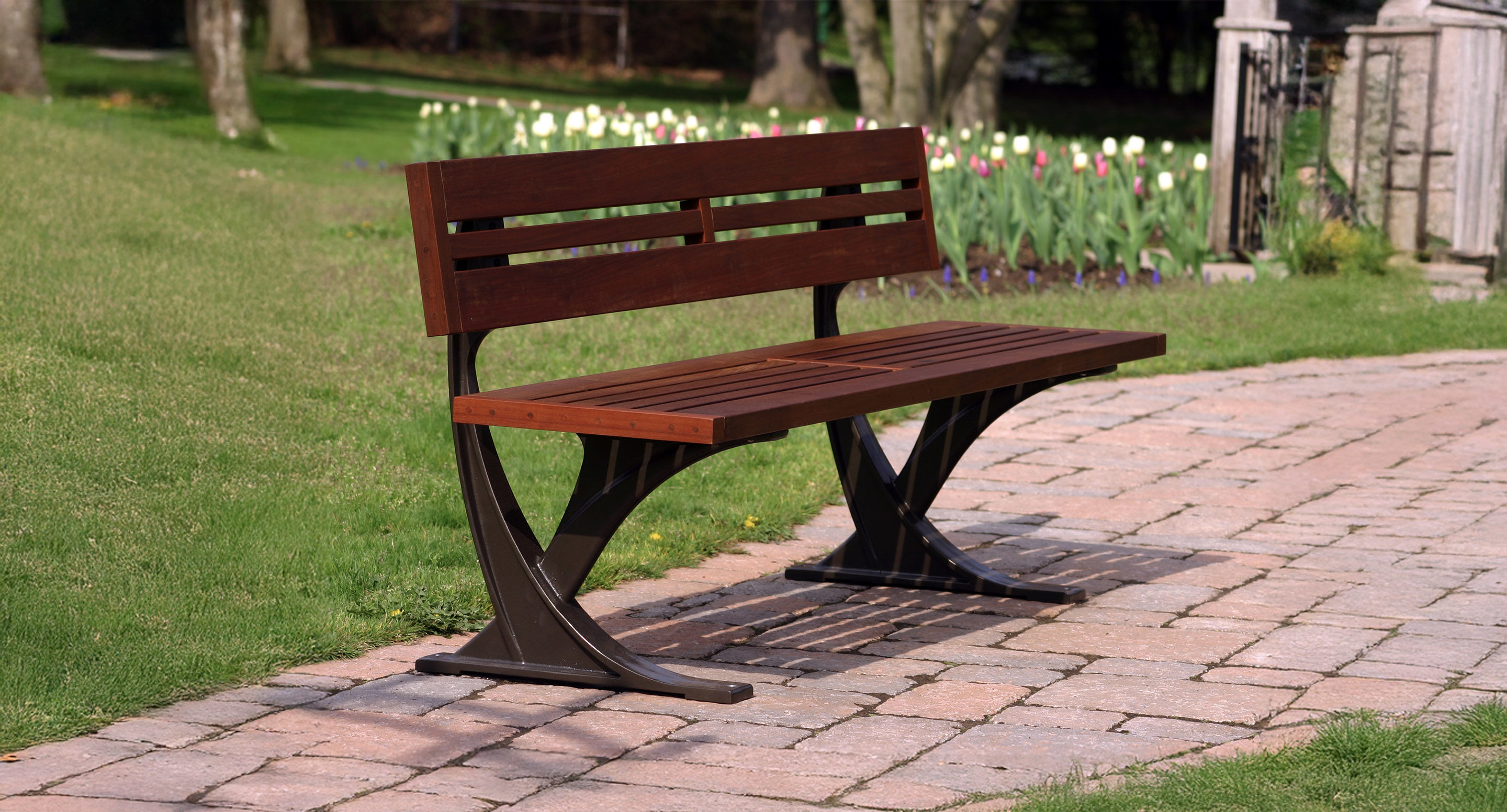 Un banc public en bois avec un cadre en métal est placé sur un sentier en pierre dans un parc herbeux avec des arbres et des fleurs en arrière-plan.