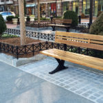 Une rangée de bancs en bois vides borde une allée pavée dans une zone urbaine aménagée avec des arbustes, des petits arbres et des devantures de magasins en arrière-plan.