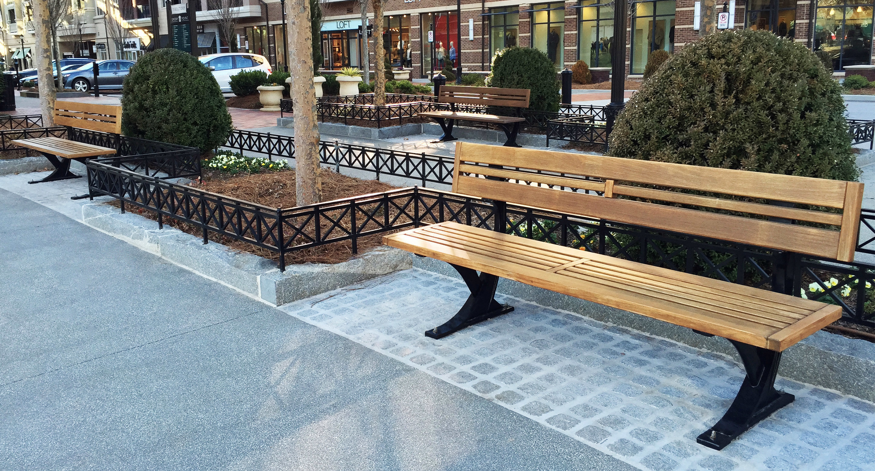 Une rangée de bancs en bois vides borde une allée pavée dans une zone urbaine aménagée avec des arbustes, des petits arbres et des devantures de magasins en arrière-plan.