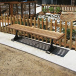 A wooden bench with metal legs is positioned on a curved concrete path next to a wooden fence in a landscaped outdoor area with rocks and plants.