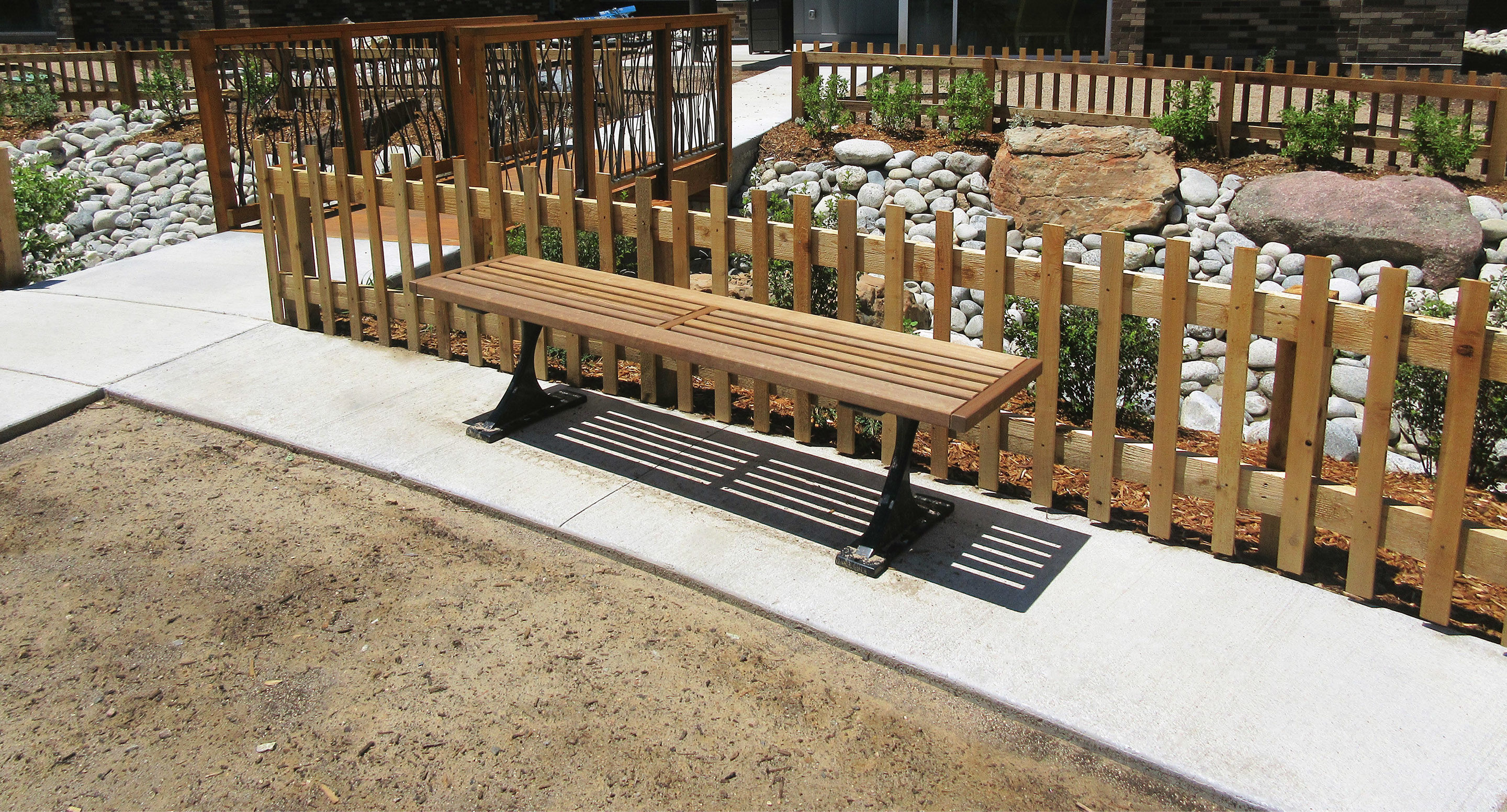 A wooden bench with metal legs is positioned on a curved concrete path next to a wooden fence in a landscaped outdoor area with rocks and plants.