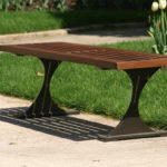 A wooden slatted bench with metal legs sits on a concrete path next to green grass and blooming tulips in a garden.