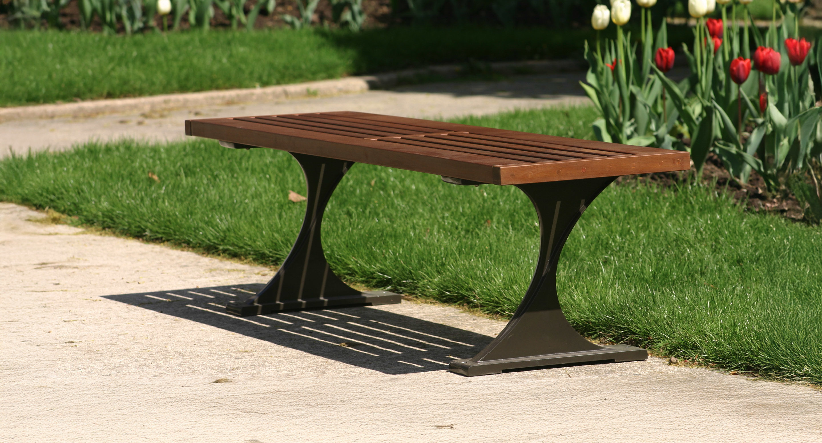 A wooden slatted bench with metal legs sits on a concrete path next to green grass and blooming tulips in a garden.
