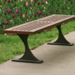 A wooden slat bench with metal legs is placed next to a sidewalk, with blooming tulips and green grass in the background.