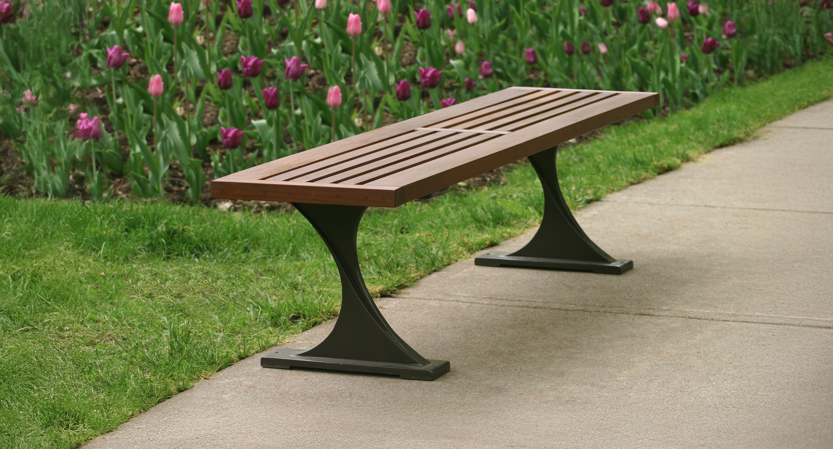 A wooden slat bench with metal legs is placed next to a sidewalk, with blooming tulips and green grass in the background.