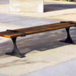 A wooden bench with metal supports is placed on a paved outdoor area in sunlight, near a concrete block and some plants.