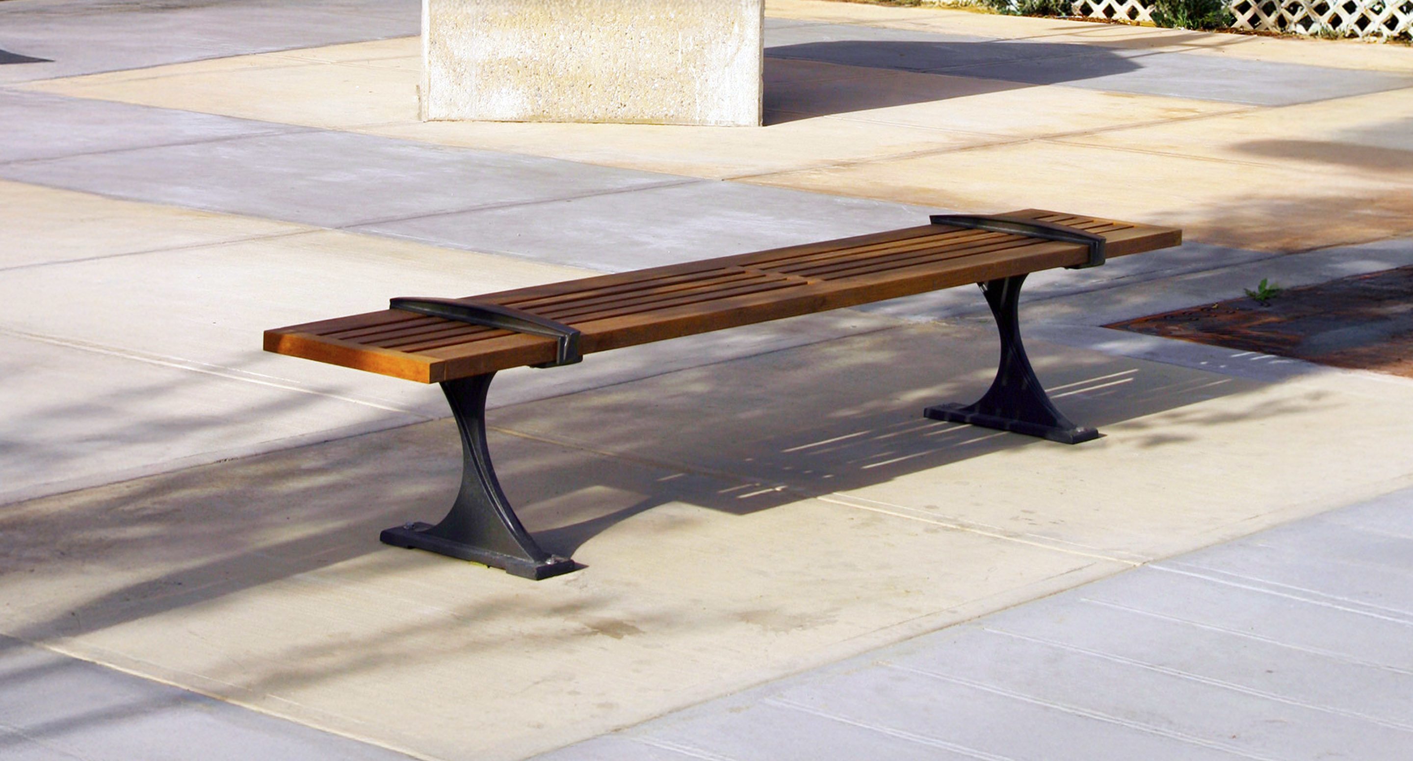 A wooden bench with metal supports is placed on a paved outdoor area in sunlight, near a concrete block and some plants.