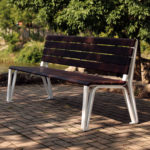 A wooden park bench with metal legs sits on a paved area near a river, surrounded by greenery and trees in the background.