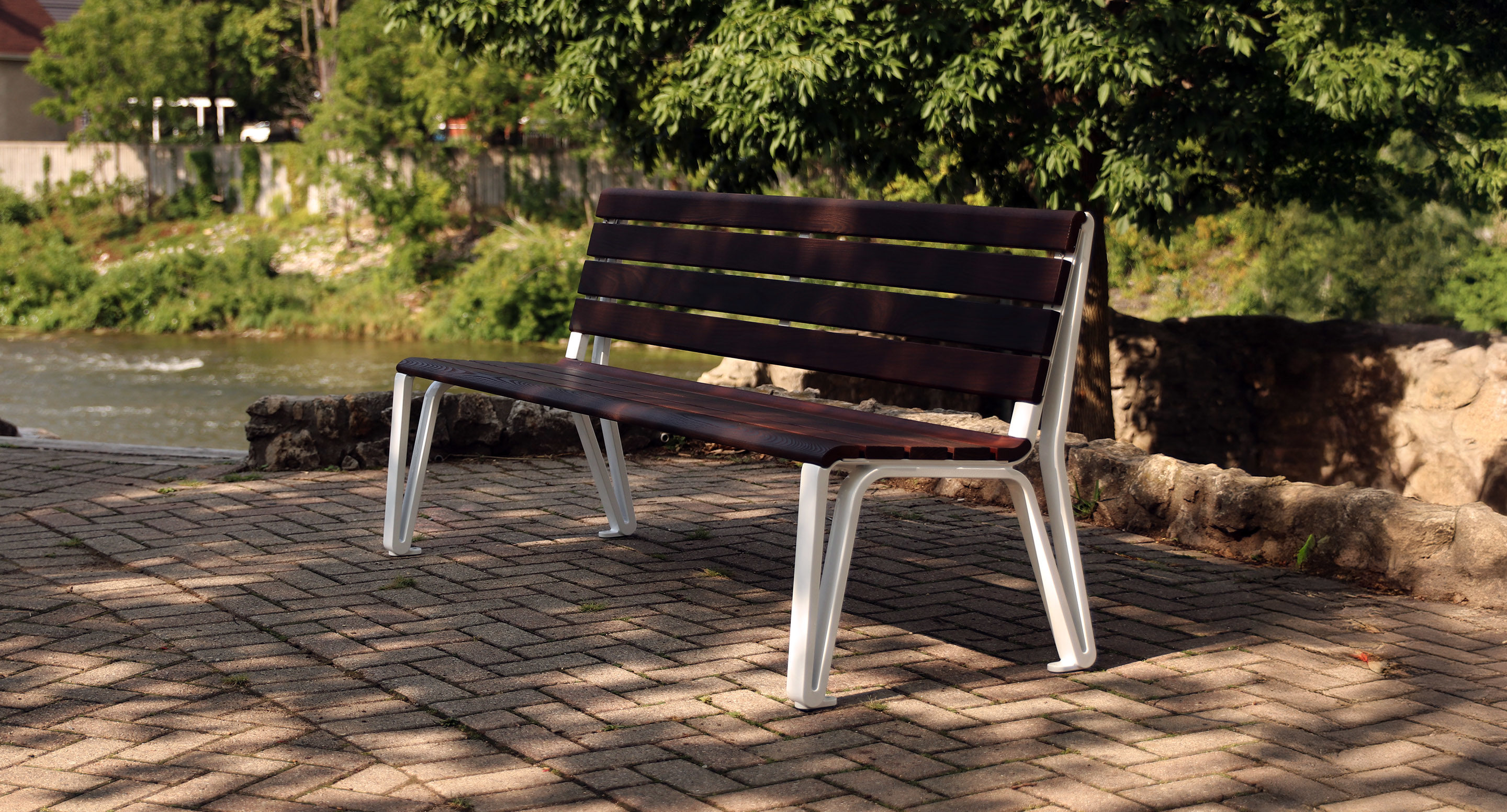 A wooden park bench with metal legs sits on a paved area near a river, surrounded by greenery and trees in the background.