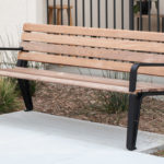 A wooden bench with black metal legs and armrests is placed on a concrete path near a landscaped area with grass and a black metal fence in the background.