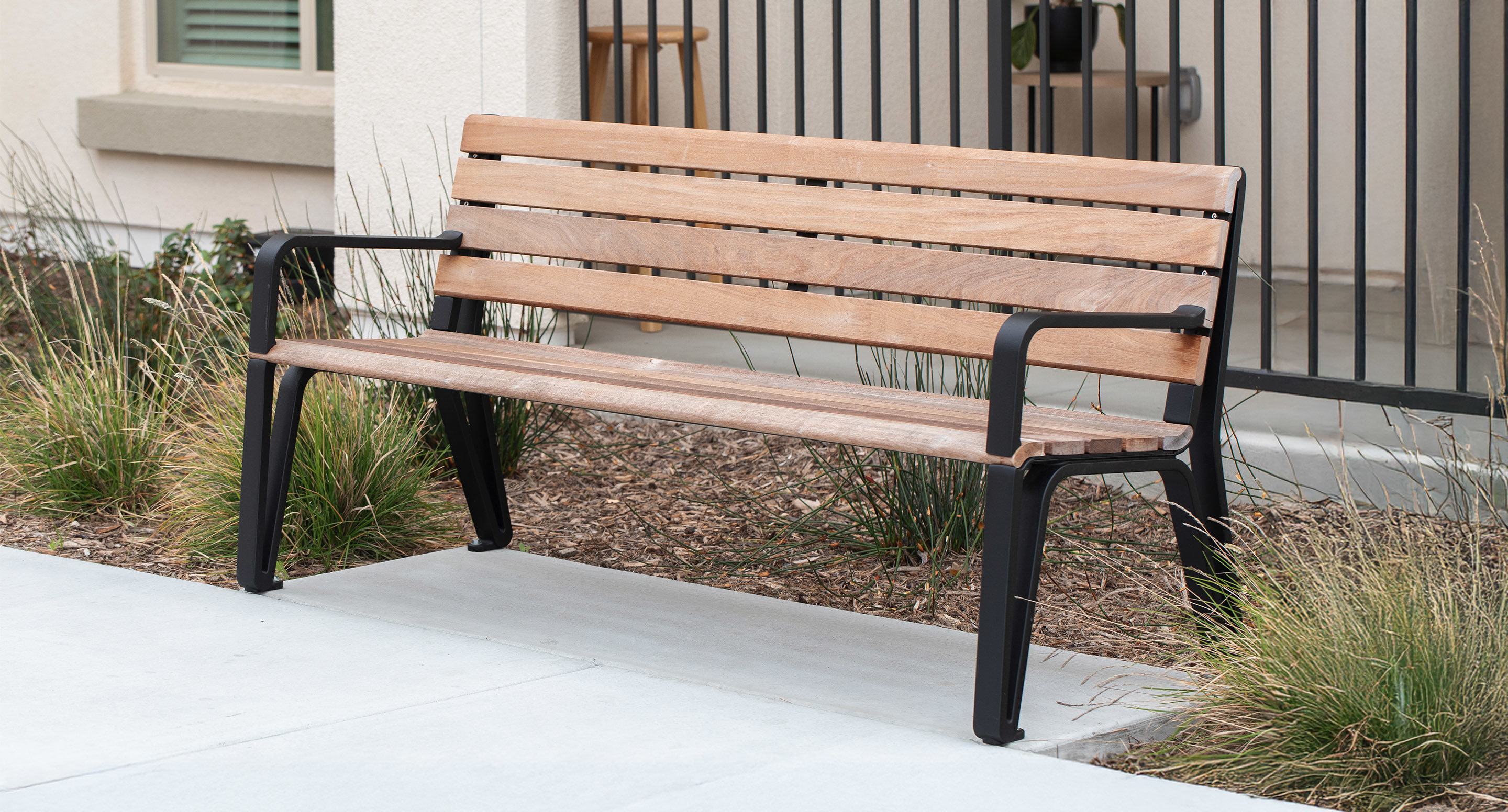 A wooden bench with black metal legs and armrests is placed on a concrete path near a landscaped area with grass and a black metal fence in the background.