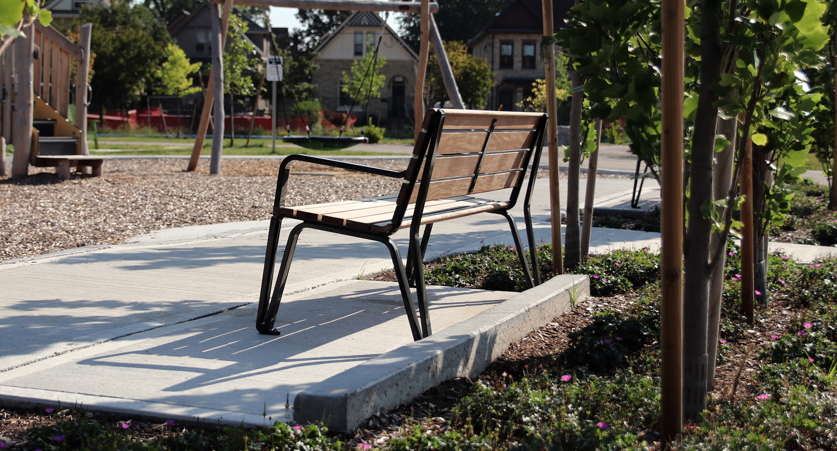 A wooden park bench sits on a paved walkway surrounded by young trees and landscaping, with houses visible in the background.