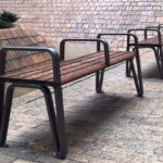 Two metal and wood benches stand on a tiled surface near a wall, with a small flower bed visible in the background.