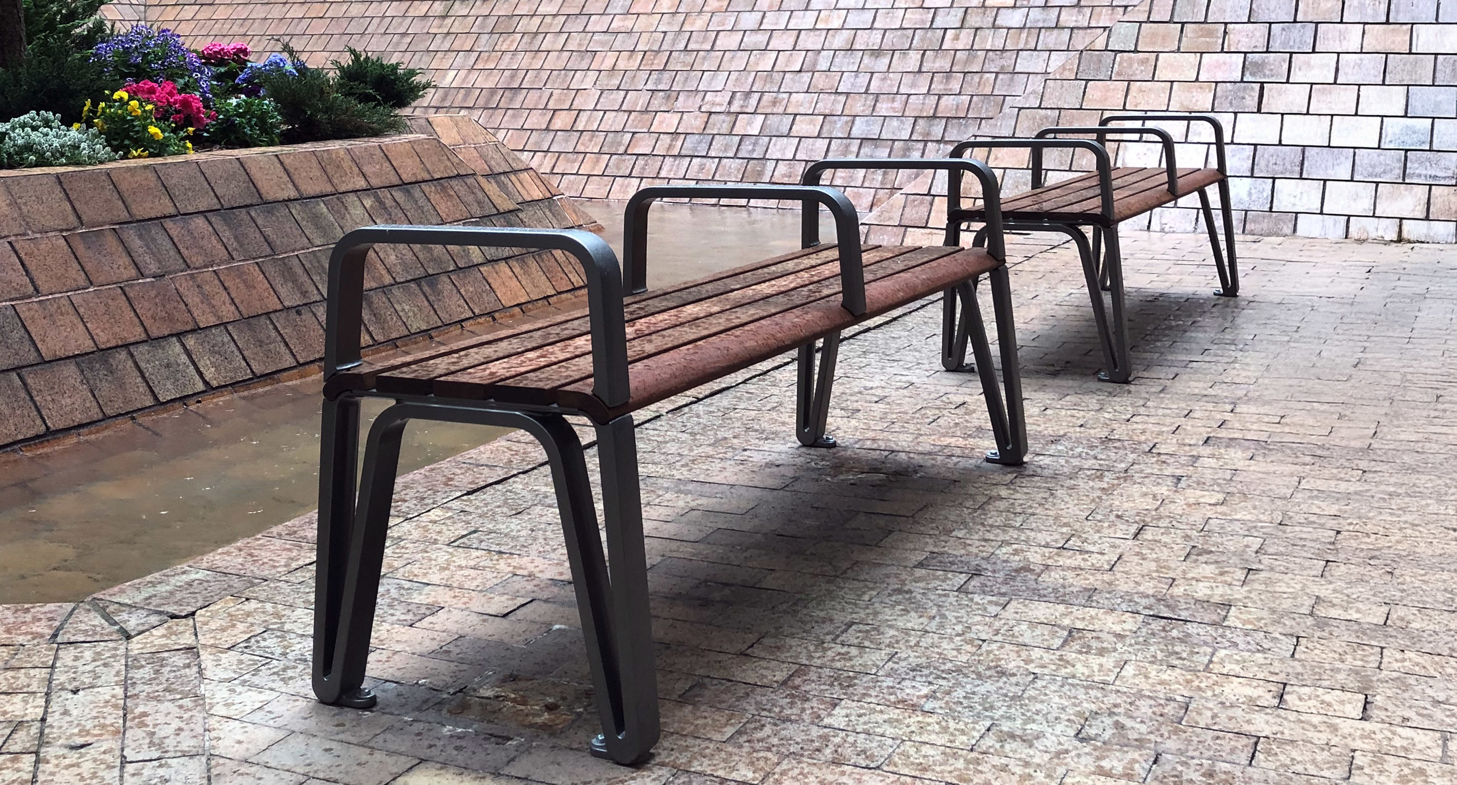 maglin_mbe-2300-backless-benches-1 Two metal and wood benches stand on a tiled surface near a wall, with a small flower bed visible in the background.