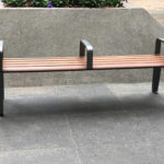 A wooden bench with metal armrests and two dividing bars is placed on a paved area near a concrete structure with steps on both sides.