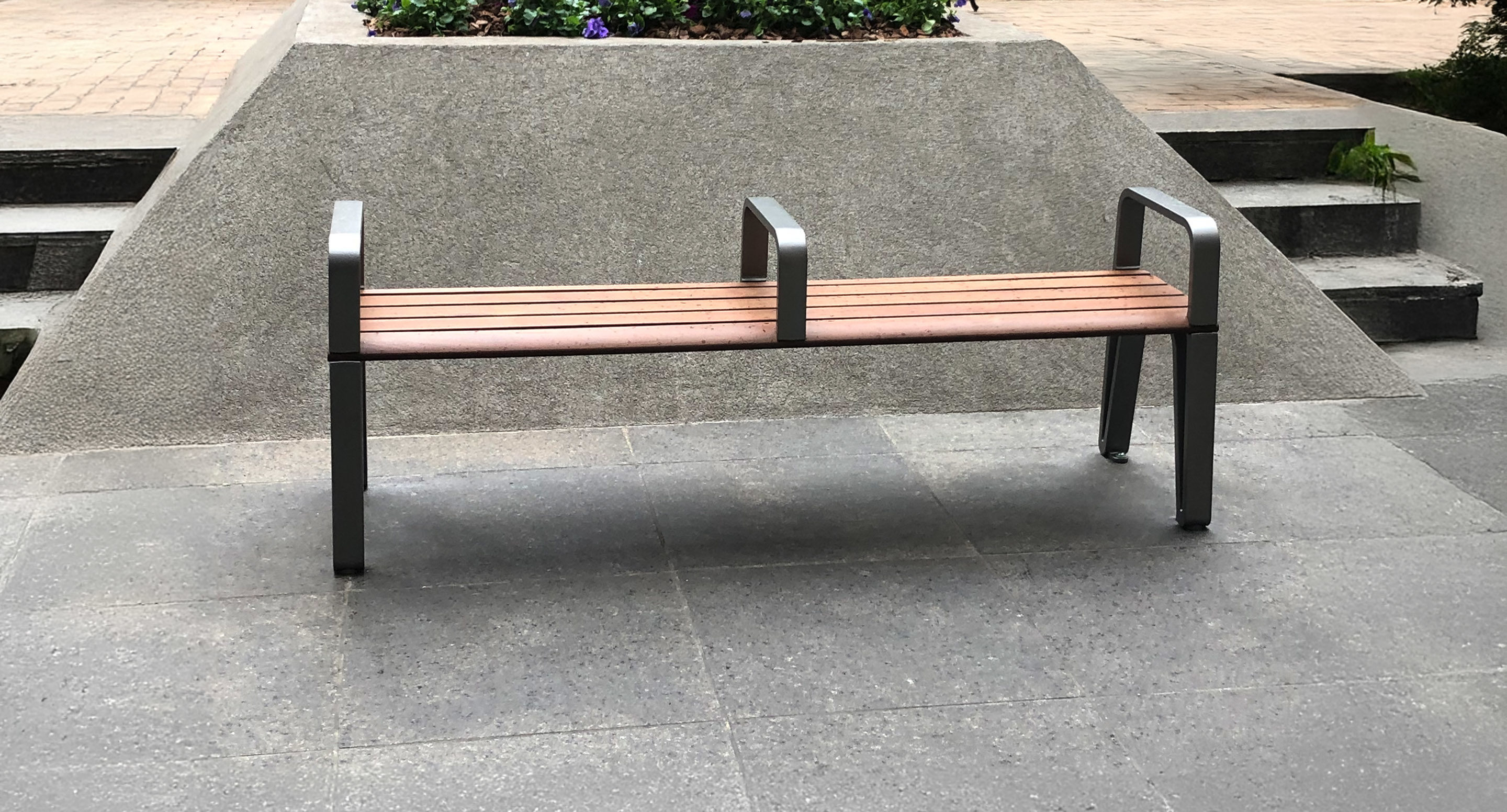 maglin_mbe-2300-backless-benches-2 A wooden bench with metal armrests and two dividing bars is placed on a paved area near a concrete structure with steps on both sides.
