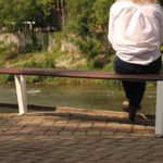 A person sits alone on a long wooden bench facing a river, with greenery and houses visible in the background.