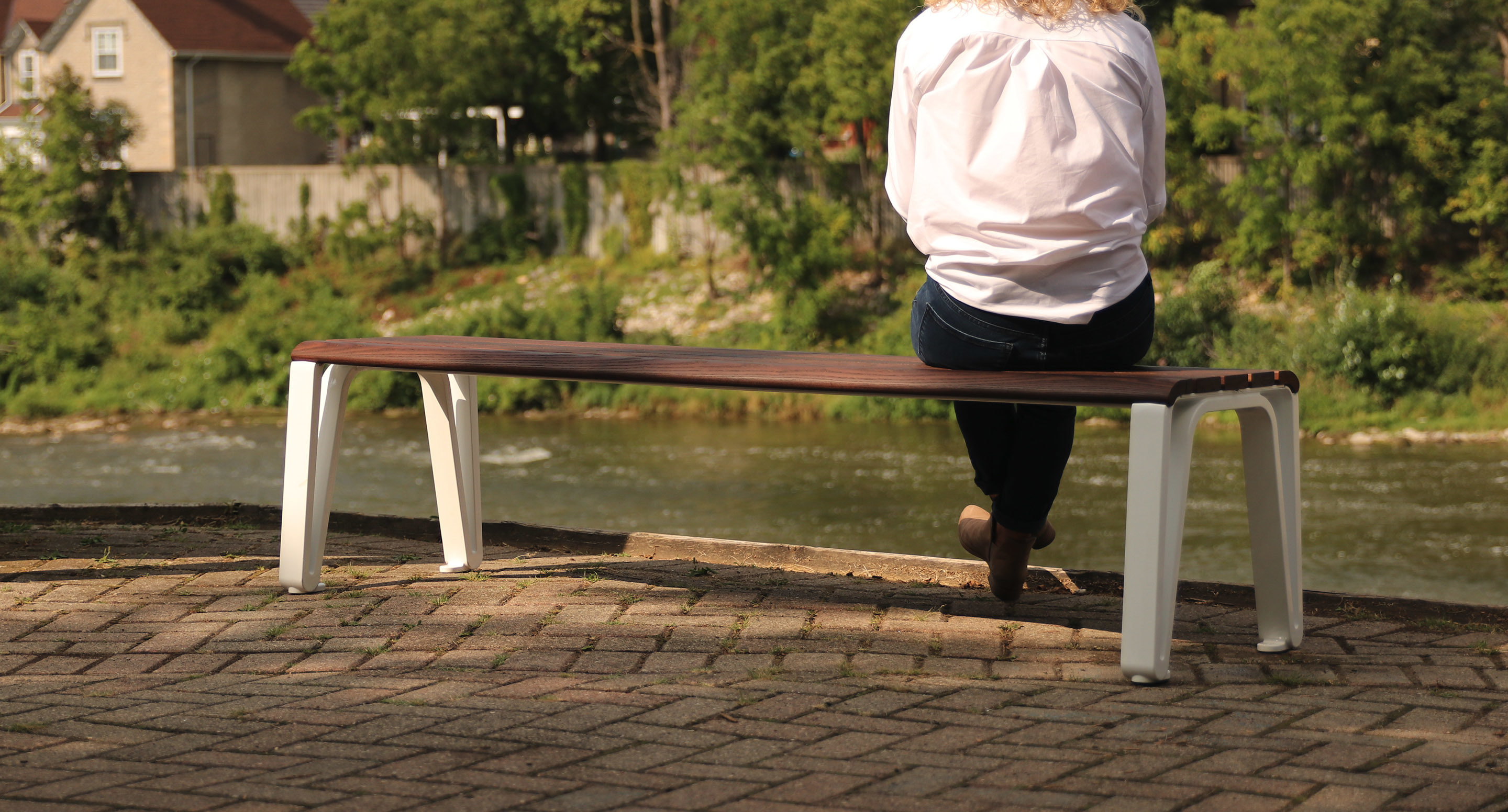 maglin_mbe-2300-backless-benches-3 A person sits alone on a long wooden bench facing a river, with greenery and houses visible in the background.