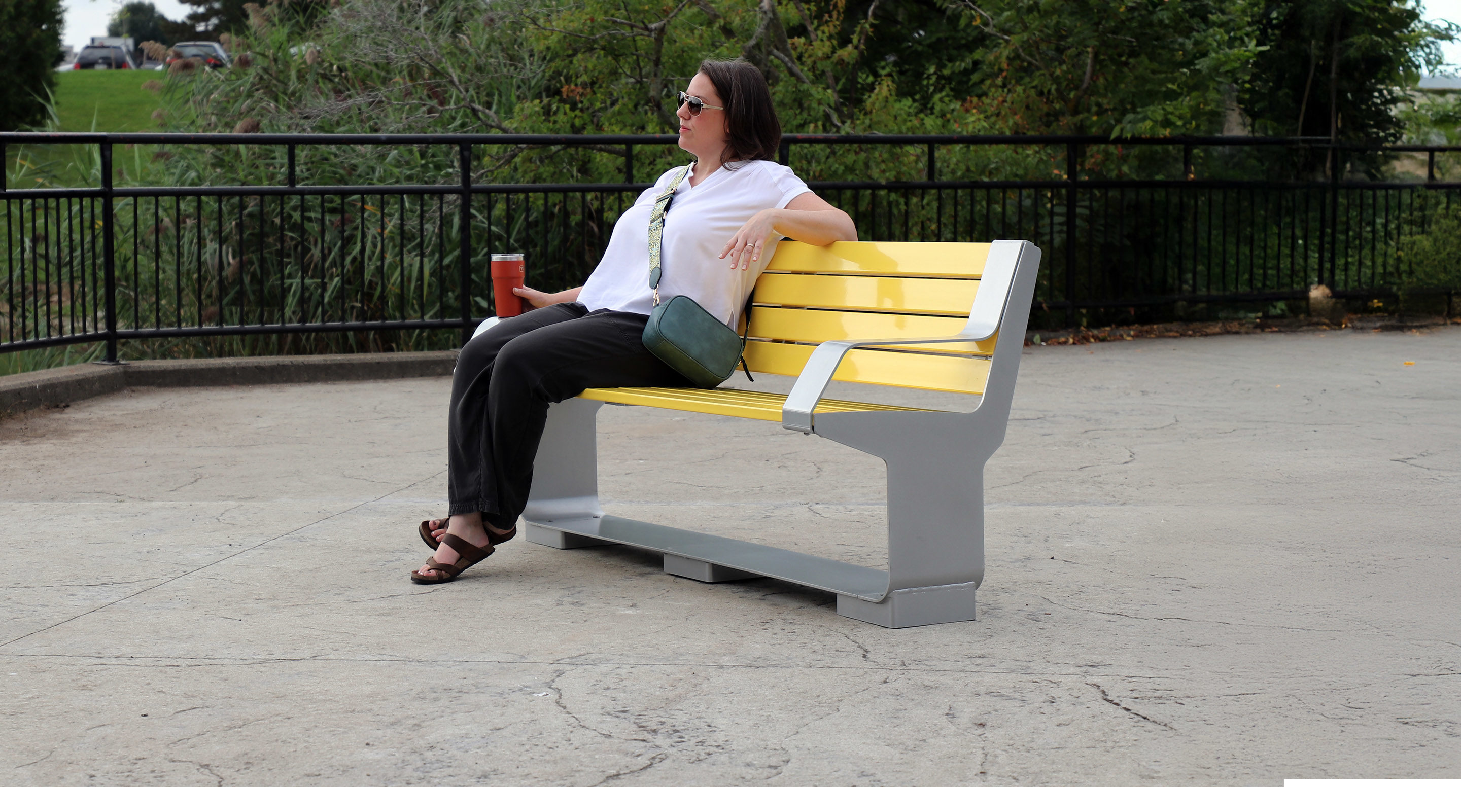 maglin_mbe-3000-backed-benches-1 A woman wearing sunglasses sits on a yellow bench outdoors, holding a drink in her hand, with trees and a black metal fence in the background.
